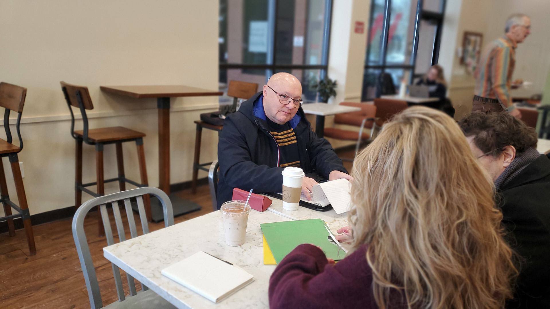 A bald man sits in a cafe, intently listening to a man and woman sitting across from him, folders and cups adorn the table.